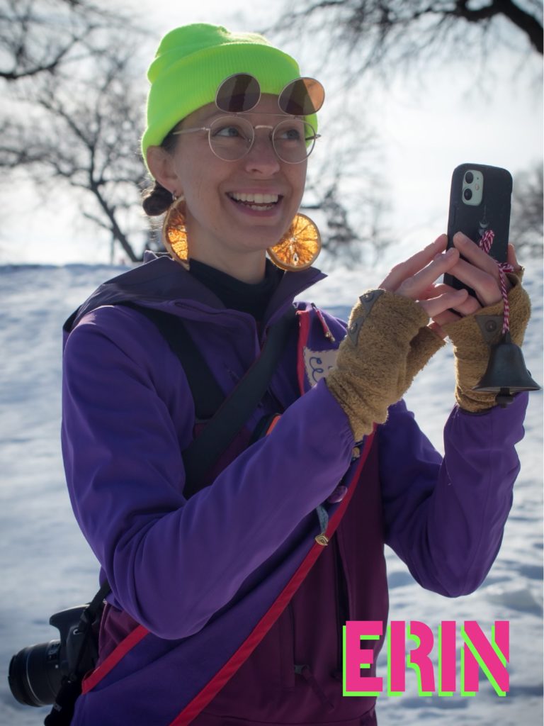 A person in brightly colored winterwear, orange slice earrings and flip up sunglasses takes a photo with their phone. Their name, "Erin" is stenciled at the bottom