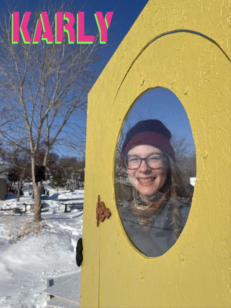 A person peers out of a round window cut out of a yellow shanty door. Their name, "Karly" is stenciled above