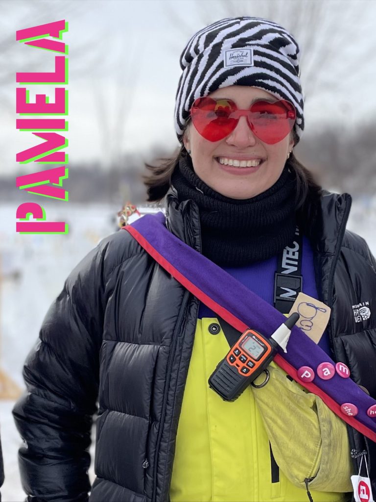 A person wearing a zebra striped hat and red tinted heart shaped glasses smiles in their uniform of a sash and walkie talkie. Their name, "Pamela" is stenciled vertically across the side