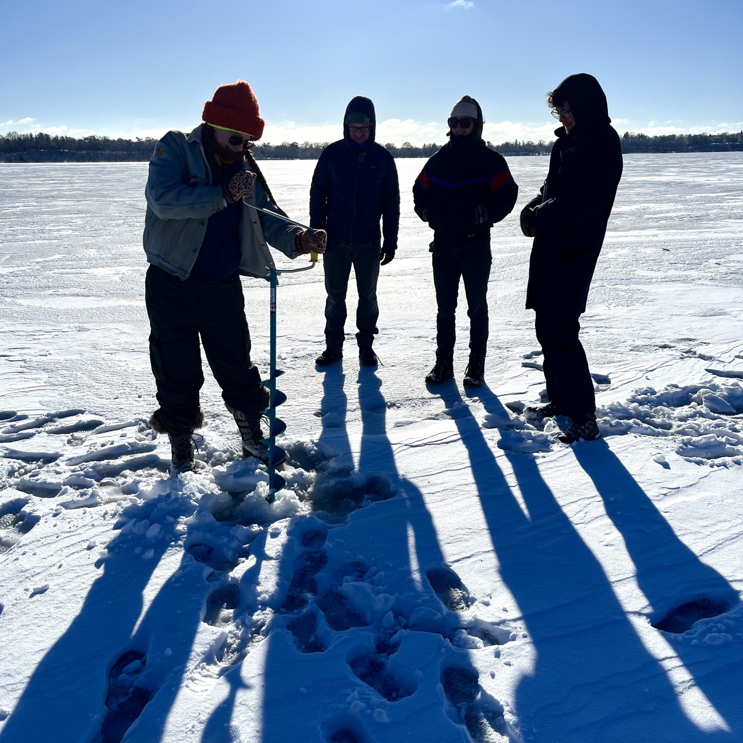 Four people in silhouette on the frozen lake. One uses a manual auger to drill a hole for an ice measurement
