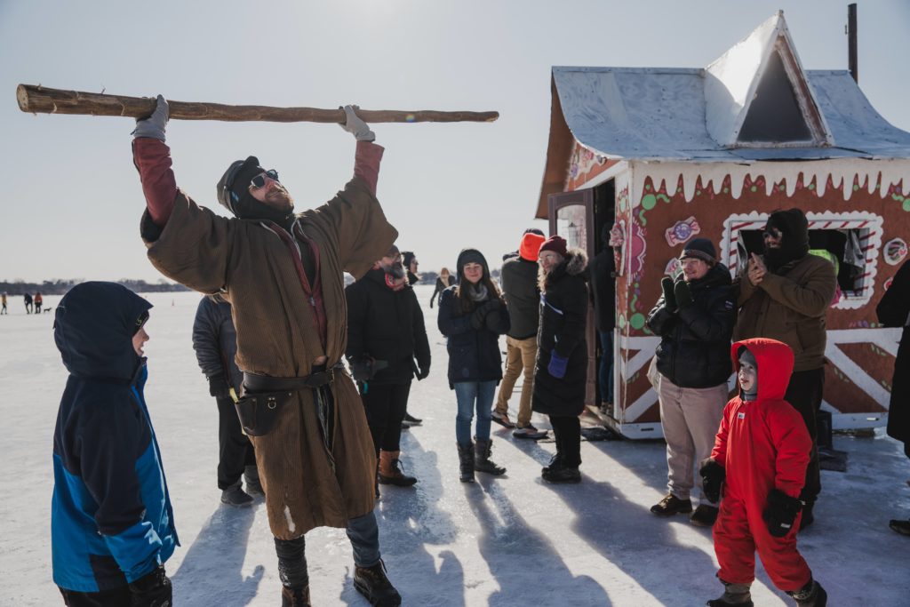A character holds a giant wooden staff above his head outside a gingerbread house shanty