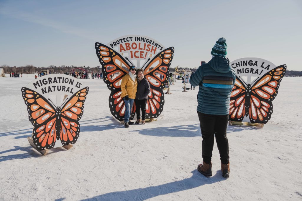 A person takes a picture of two people standing in front of a giant monarch butterfly mural on the frozen lake