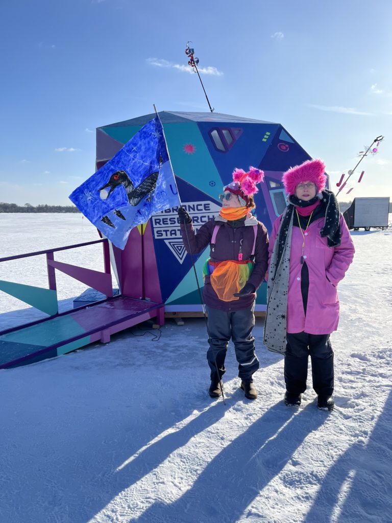 Two aliens stand outside the vibrance research station, one holding a loon flag