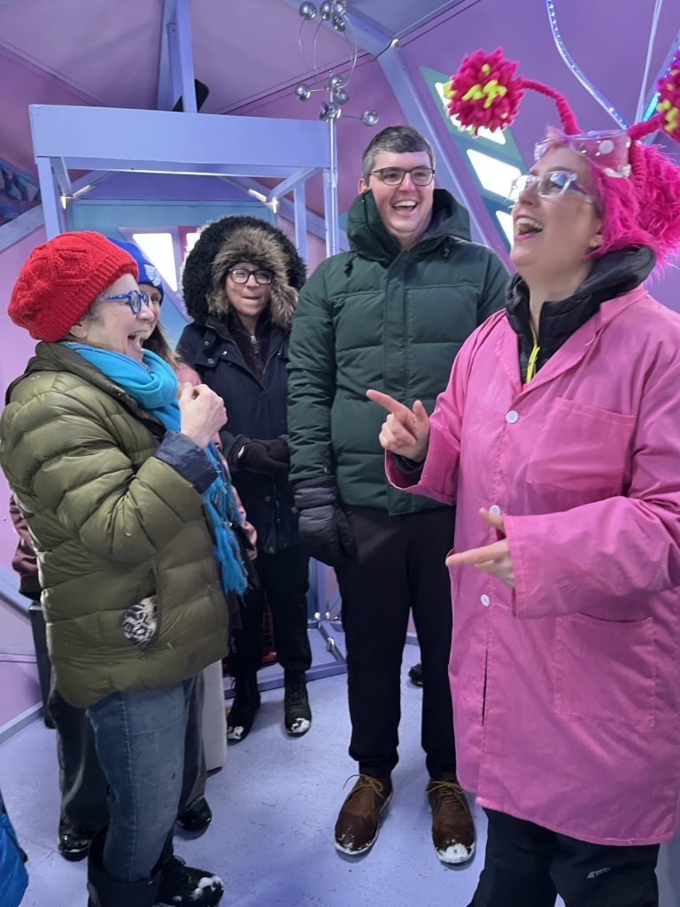 An alien character with antenna and pink hair throws their head back in laughter while addressing a crowd inside a purple shanty