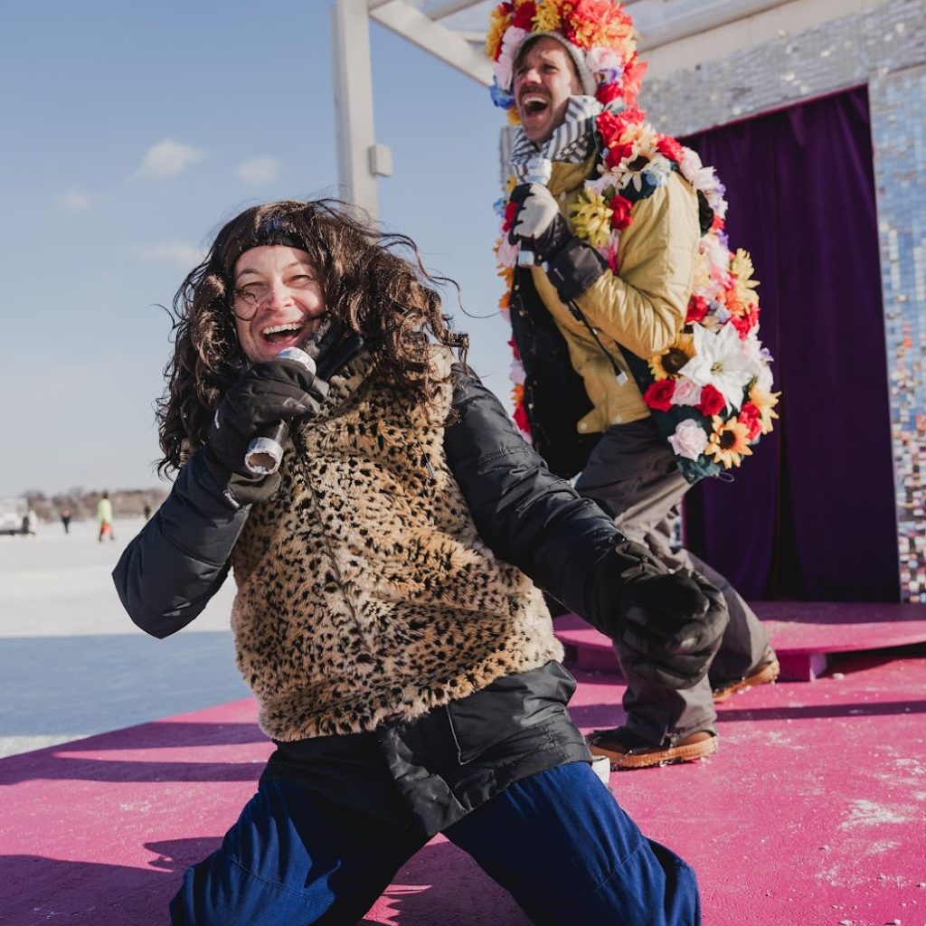 two people wear wild costume pieces over their snowsuits and perform on a pink stage
