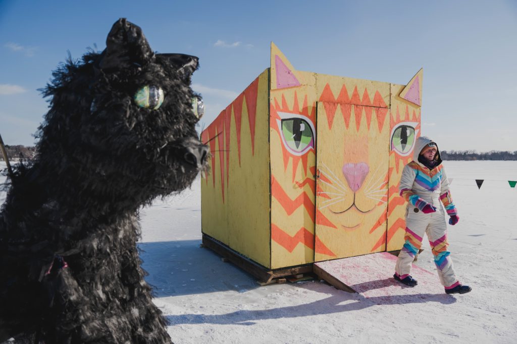 an artist in a colorful snowsuit walks out of a cat shaped shanty. a giant black cat puppet looks on