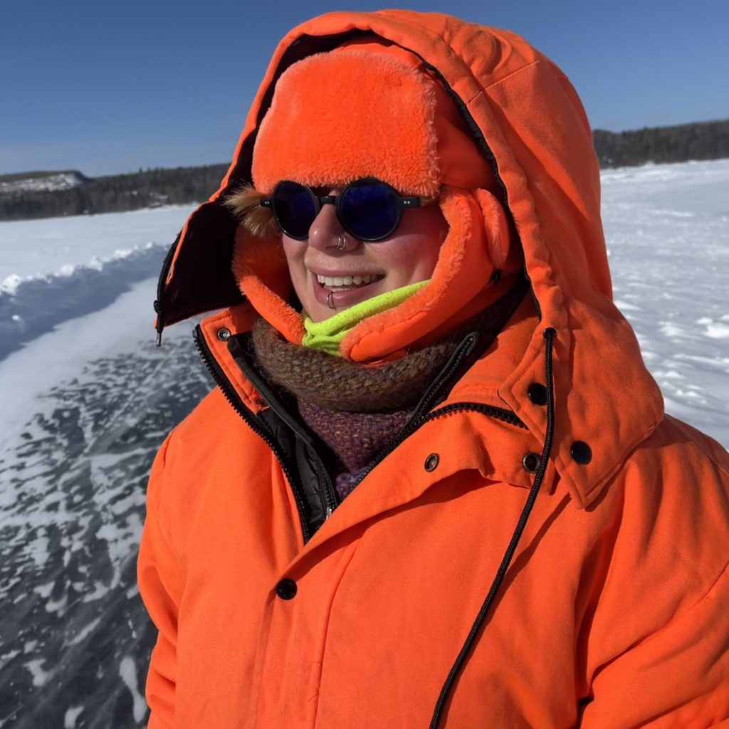 A person bundled up in blaze orange winterwear and sunglasses smiles broadly while standing on a frozen lake