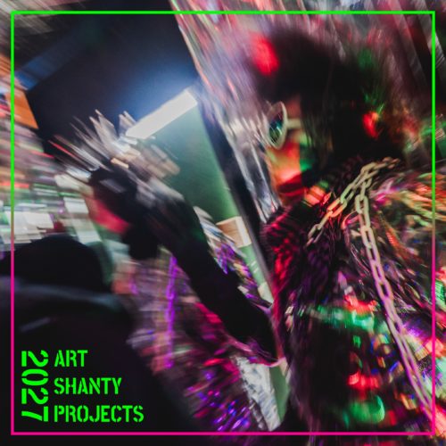 A blurry scene of two people inside a shanty with silver and vibrant color scattered by light. Neon green stencil font reads "2027 ART SHANTY PROJECTS"