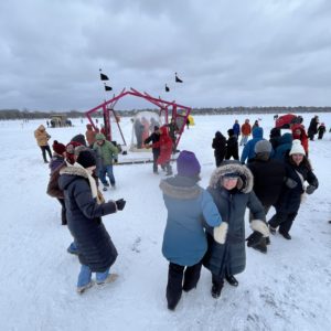 A group of people in snowsuits square dance on a frozen lake with a stage and live musicians in the distance