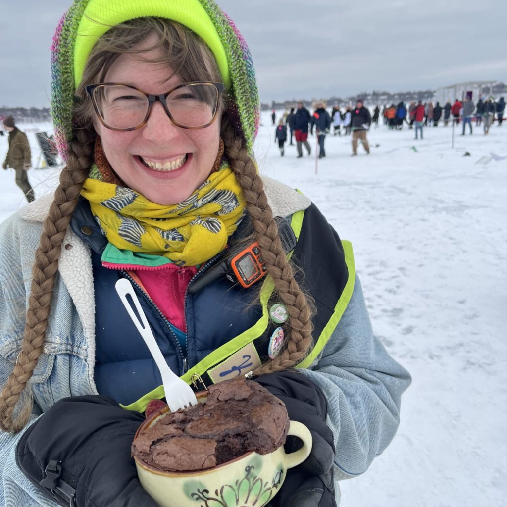 a person bundled up with long braided hair smiles while holding a mug of cake in the shanty village