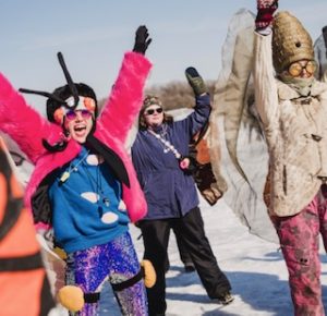 A group of adults in pollinator costumes over their snowsuits dance in the shanty village