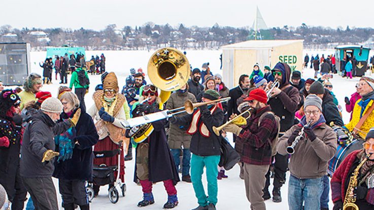 a large crowd in colorful winter-wear gathers around musicians on the frozen lake, with shanties and people in the background