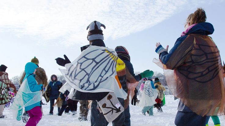 A crowd of people dance around in butterfly wings on a snow-covered lake